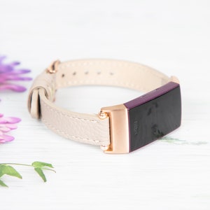 Puede incluir: Una banda de cuero blanco con detalles en oro rosa para un rastreador de actividad f&iacute;sica Fitbit. La banda se muestra sobre una superficie blanca.