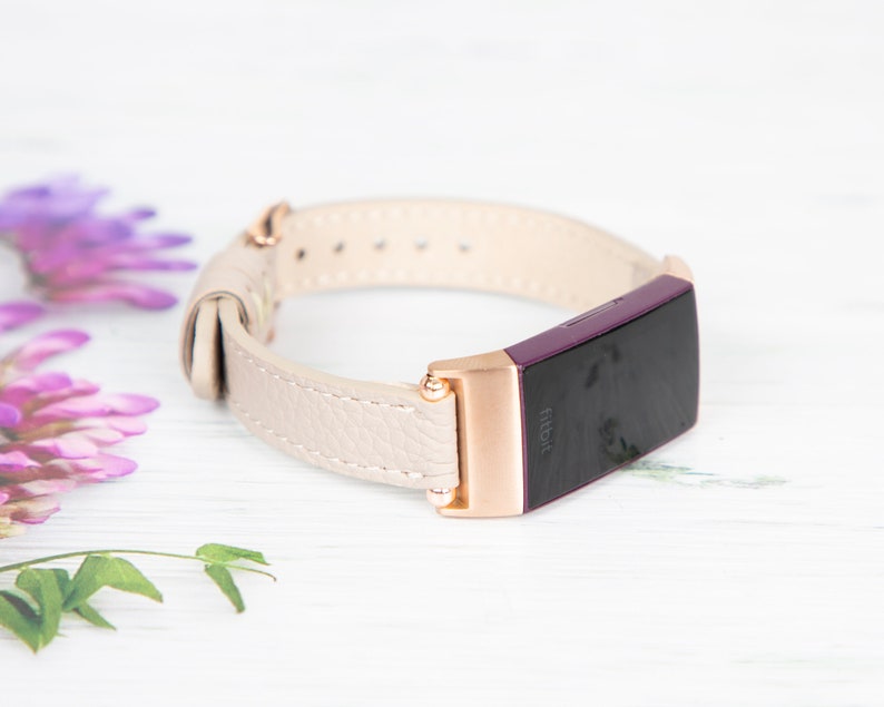 Puede incluir: Una banda de cuero blanco con detalles en oro rosa para un rastreador de actividad f&iacute;sica Fitbit. La banda se muestra sobre una superficie blanca.