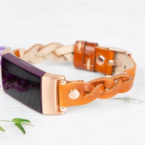 May include: A braided leather band for a fitness tracker in brown and white with a rose gold buckle. The band is shown on a purple fitness tracker.