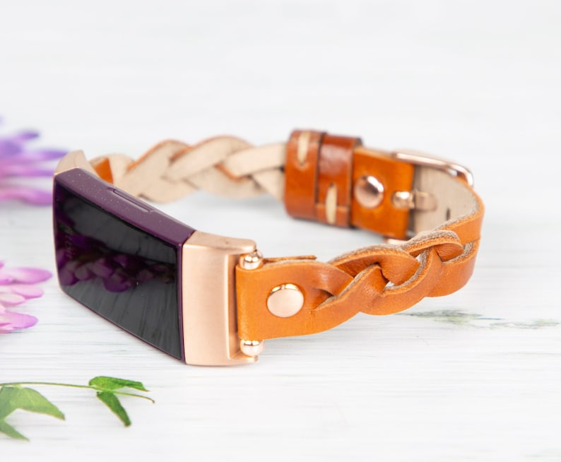 May include: A braided leather band for a fitness tracker in brown and white with a rose gold buckle. The band is shown on a purple fitness tracker.