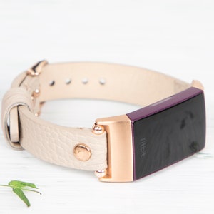 Puede incluir: Una banda de cuero crema con detalles en oro rosa y un rastreador Fitbit morado. El rastreador tiene la palabra "Fitbit" impresa en la pantalla.