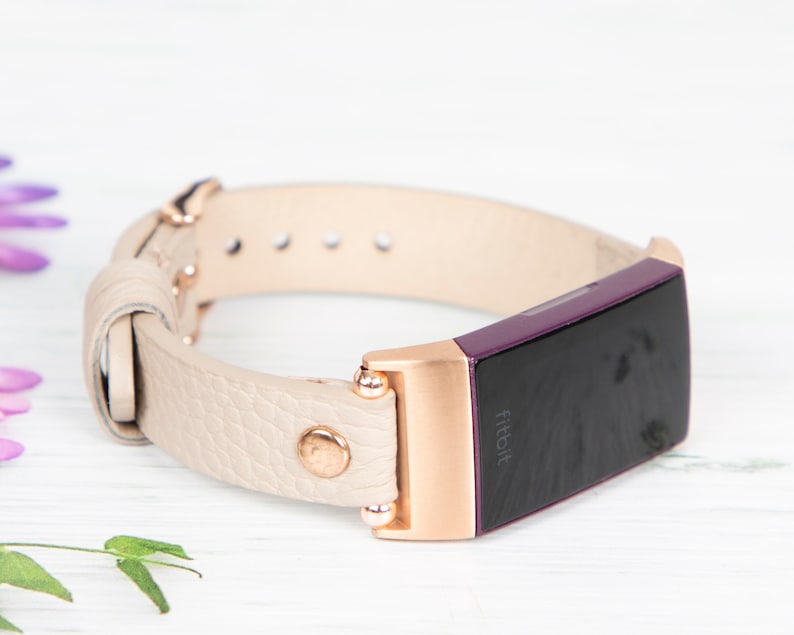 Puede incluir: Una banda de cuero crema con detalles en oro rosa y un rastreador Fitbit morado. El rastreador tiene la palabra "Fitbit" impresa en la pantalla.