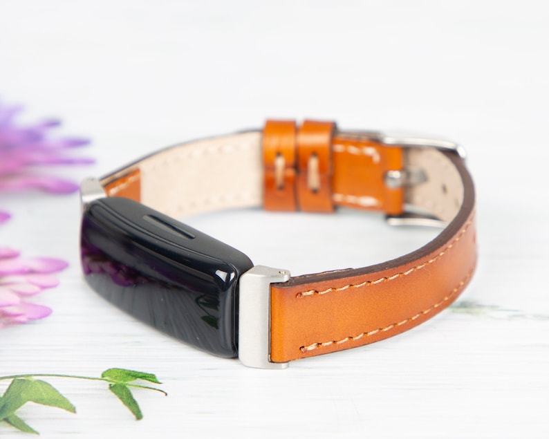 May include: A black fitness tracker with a brown leather band. The band has white stitching and a silver buckle.