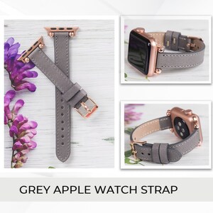 May include: A grey leather Apple Watch band with rose gold hardware. The band is shown on a white wooden surface with purple flowers.