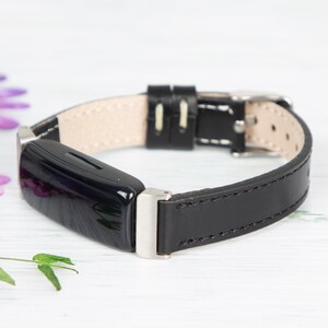 May include: A black leather band with a silver buckle and a black fitness tracker. The band has white stitching.