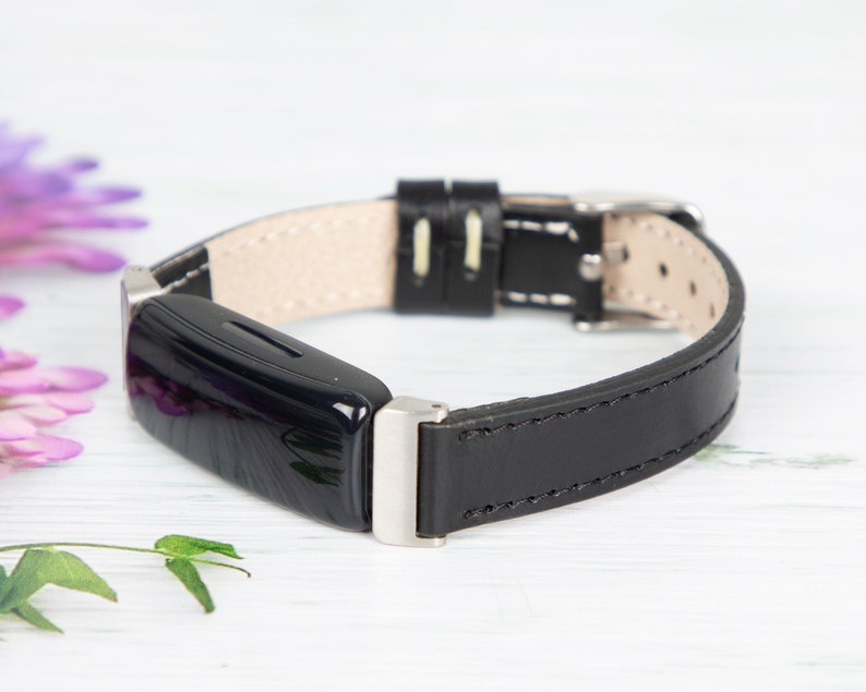May include: A black leather band with a silver buckle and a black fitness tracker. The band has white stitching.