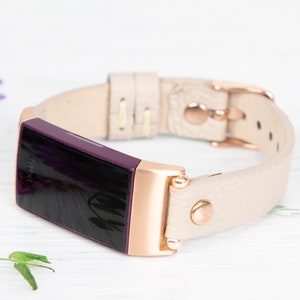 Puede incluir: Un rastreador de actividad f&iacute;sica Fitbit Inspire 2 de color rosa dorado y morado con una banda de cuero crema. La banda tiene una hebilla y costuras de color rosa dorado.
