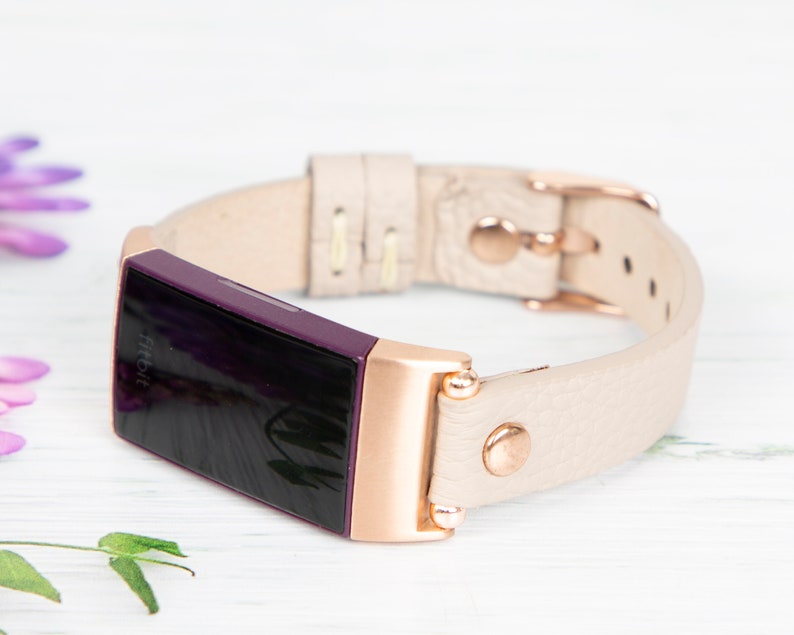 Puede incluir: Un rastreador de actividad f&iacute;sica Fitbit Inspire 2 de color rosa dorado y morado con una banda de cuero crema. La banda tiene una hebilla y costuras de color rosa dorado.