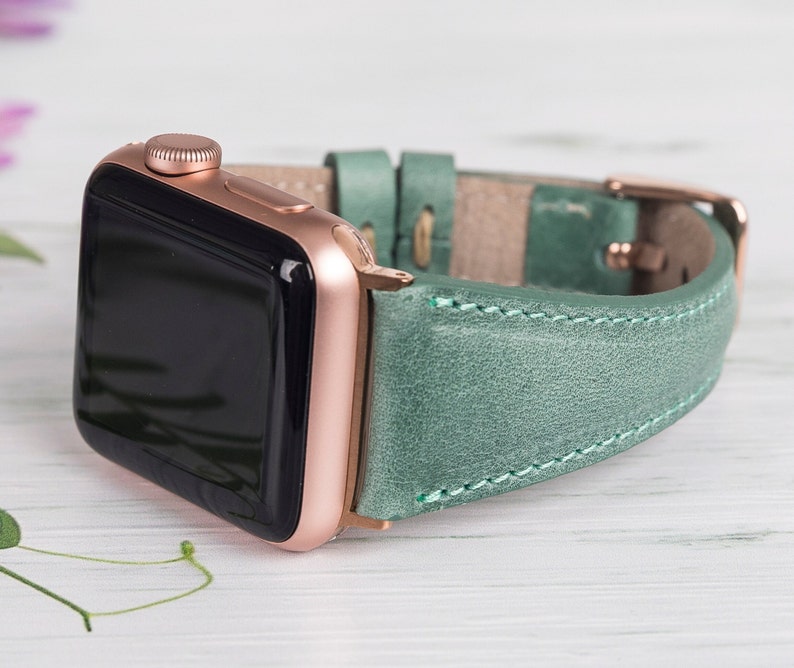 Puede incluir: Un Apple Watch de oro rosa con una correa de cuero turquesa. La correa tiene costuras blancas y una hebilla de oro rosa.