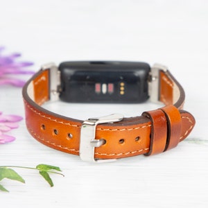 May include: A brown leather watch strap with a silver buckle. The strap is designed to fit a fitness tracker.