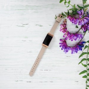 Puede incluir: Una pulsera de cuero beige con una esfera de reloj digital negra sobre un fondo de madera blanca con flores moradas.
