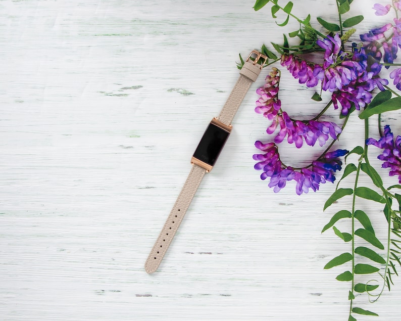 Puede incluir: Una pulsera de cuero beige con una esfera de reloj digital negra sobre un fondo de madera blanca con flores moradas.