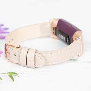 Puede incluir: Una banda de cuero beige claro con una hebilla de oro rosa y un rastreador de fitness morado.