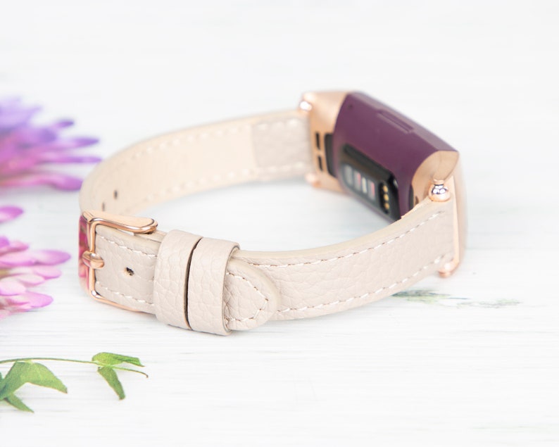 Puede incluir: Una banda de cuero beige claro con una hebilla de oro rosa y un rastreador de fitness morado.
