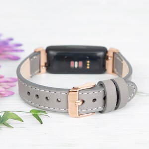May include: A grey leather band with rose gold hardware for a fitness tracker. The band has a buckle closure and is designed to fit a small wrist.
