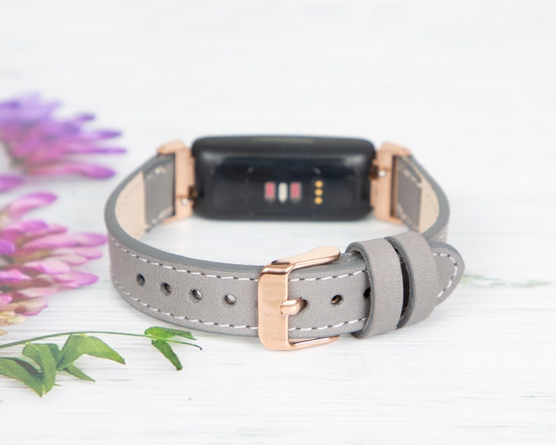May include: A grey leather band with rose gold hardware for a fitness tracker. The band has a buckle closure and is designed to fit a small wrist.