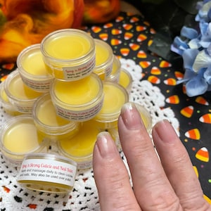 May include: A close-up of a small jar of cuticle and nail balm. The jar is labelled "Long 'n Strong Cuticle and Nail Balm". Apply and massage daily to clean cuticles and nails. May also be used over polish.