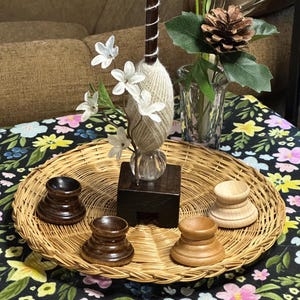 May include: Four wooden candle holders, two brown and two natural wood, are arranged on a woven wicker tray. The tray is on a black and white floral tablecloth. A small wooden pedestal with a glass vase containing a white yarn ball and white flowers is in the center of the tray.