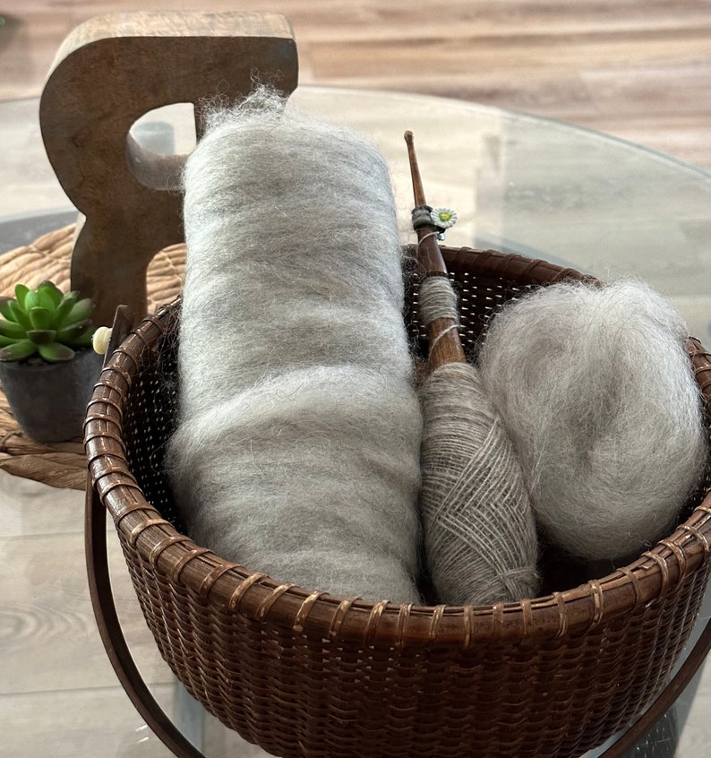 Puede incluir: Una cesta de mimbre llena de tres ovillos de lana gris claro. Un ovillo est&aacute; enrollado alrededor de un huso de madera con un amuleto de flor blanca.