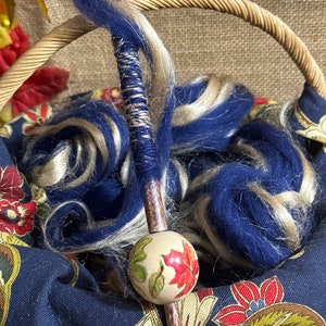 May include: A wooden spinning spindle with a decorative, hand-painted bead featuring autumn leaves. The spindle is wrapped in blue and white yarn and is resting in a basket.