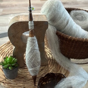 Puede incluir: Un torno de hilar de madera con un huso de hilo gris y una madeja de hilo gris en una cesta de mimbre. El hilo se est&aacute; hilando en el torno y una peque&ntilde;a planta est&aacute; en una maceta sobre la mesa.