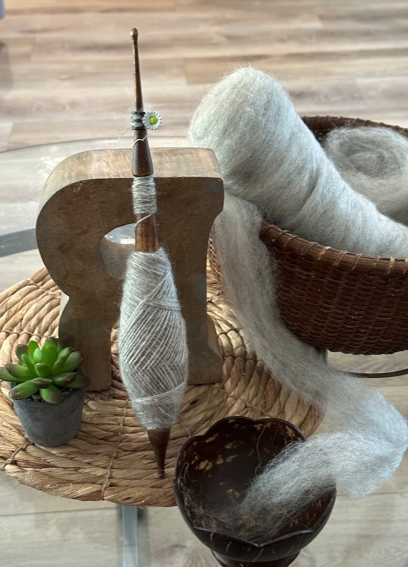 Puede incluir: Un torno de hilar de madera con un huso de hilo gris y una madeja de hilo gris en una cesta de mimbre. El hilo se est&aacute; hilando en el torno y una peque&ntilde;a planta est&aacute; en una maceta sobre la mesa.