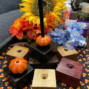 Puede incluir: Un conjunto de seis portavelas de madera, cada uno con un pequeño pomo en forma de calabaza en la parte superior. Los portavelas están teñidos en varios colores, incluyendo negro, marrón y rojo. Los portavelas están dispuestos sobre una superficie con un patrón blanco y negro.