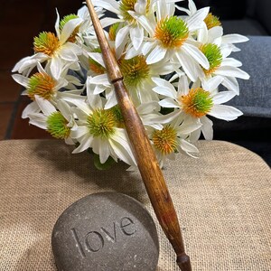 Puede incluir: Un huso de madera con un patr&oacute;n de veta de madera marr&oacute;n y naranja, descansando sobre una superficie de arpillera junto a una piedra gris con la palabra "love" tallada en ella. Flores blancas con centros verdes est&aacute;n en el fondo.