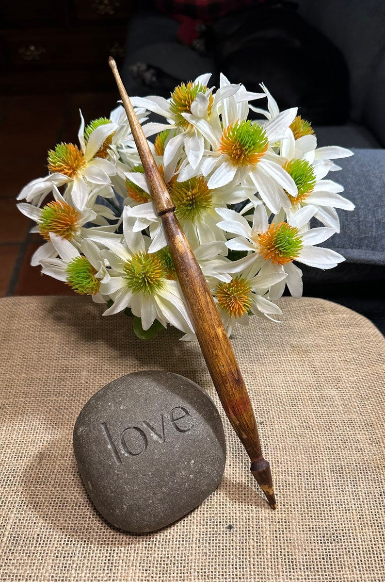 Puede incluir: Un huso de madera con un patr&oacute;n de veta de madera marr&oacute;n y naranja, descansando sobre una superficie de arpillera junto a una piedra gris con la palabra "love" tallada en ella. Flores blancas con centros verdes est&aacute;n en el fondo.