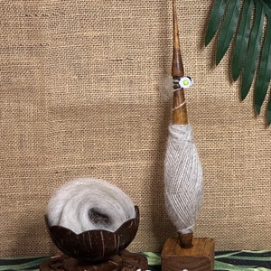 Puede incluir: Una rueca de madera con hilo de lana gris sobre ella. La rueca est&aacute; sobre una base de madera y est&aacute; rodeada de flores blancas. La rueca est&aacute; sobre una mesa con un fondo de arpillera marr&oacute;n.