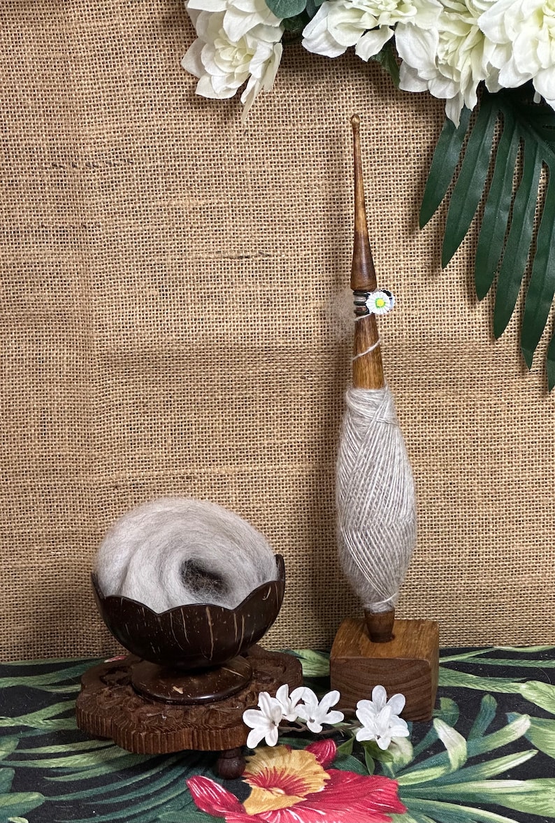 Puede incluir: Una rueca de madera con hilo de lana gris sobre ella. La rueca est&aacute; sobre una base de madera y est&aacute; rodeada de flores blancas. La rueca est&aacute; sobre una mesa con un fondo de arpillera marr&oacute;n.