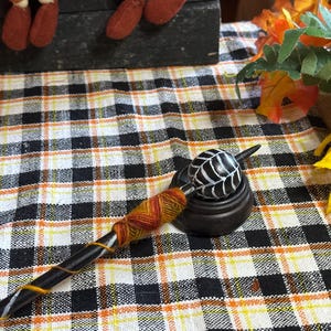 May include: A black and white spindle with a black and white ball on the end. The spindle is wrapped with orange and yellow yarn. The spindle is sitting on a black wooden base.