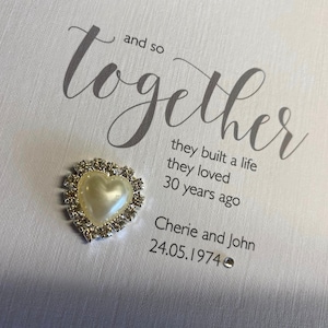 以下が含まれることがあります： 「and so together」という言葉を筆記体で書いた白いカード。その下に「they built a life they loved 30 years ago」という文章が書かれている。さらにその下に「Cherie and John 24.05.1974」という文章が書かれている。カードには、銀色の縁取りとラインストーンが付いた小さな白いハートが付けられている。