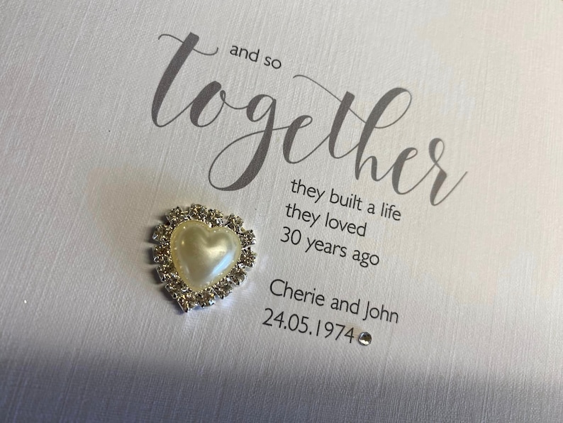 以下が含まれることがあります： 「and so together」という言葉を筆記体で書いた白いカード。その下に「they built a life they loved 30 years ago」という文章が書かれている。さらにその下に「Cherie and John 24.05.1974」という文章が書かれている。カードには、銀色の縁取りとラインストーンが付いた小さな白いハートが付けられている。