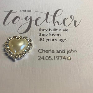 以下が含まれることがあります： 白いカードに、「and so together」という文字が筆記体で書かれています。その下に、「they built a life they loved 30 years ago」という文章があります。さらにその下に、「Cherie and John 24.05.1974」という文章があります。カードには、真珠がついた小さな銀色のハートが付けられています。