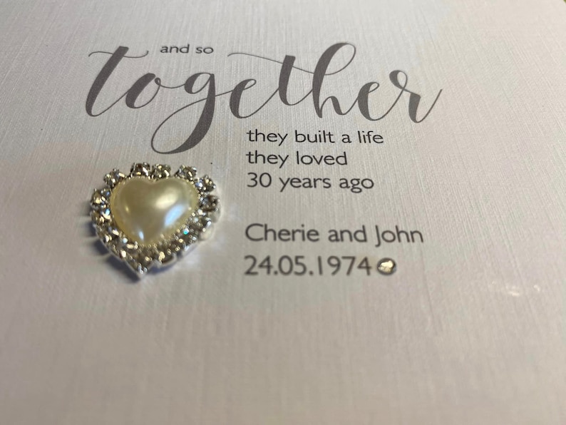 以下が含まれることがあります： 白いカードに、「and so together」という文字が筆記体で書かれています。その下に、「they built a life they loved 30 years ago」という文章があります。さらにその下に、「Cherie and John 24.05.1974」という文章があります。カードには、真珠がついた小さな銀色のハートが付けられています。