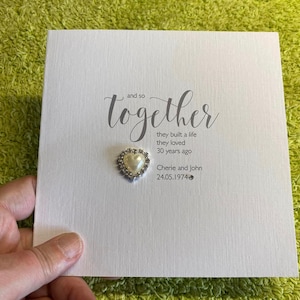 以下が含まれることがあります： グレーの筆記体で「and so together」と書かれた白いカード。テキストの下には、「they built a life they loved 30 years ago」という詩が書かれています。カードには、「Cherie and John」という名前と「24.05.1974」という日付も記載されています。カードには、真珠で飾られた小さなハートが付けられています。