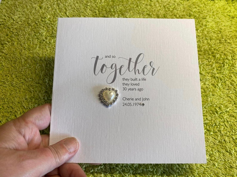 以下が含まれることがあります： グレーの筆記体で「and so together」と書かれた白いカード。テキストの下には、「they built a life they loved 30 years ago」という詩が書かれています。カードには、「Cherie and John」という名前と「24.05.1974」という日付も記載されています。カードには、真珠で飾られた小さなハートが付けられています。