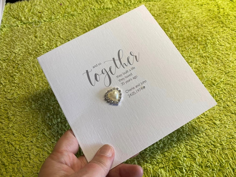 以下が含まれることがあります： 「and so together」というテキストが筆記体で書かれた白いカード。カードには、「they built a life they loved 30 years ago」と「Cherie and John 24.05.1974」というテキストも含まれています。カードには、真珠で飾られた小さなハート型の装飾が取り付けられています。
