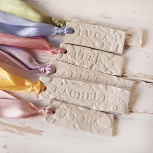 Könnte beinhalten: Nahaufnahme von mehreren rechteckigen, cremefarbenen Anhängern mit eingeprägten Namen, darunter "LUCA", "LISA", "ALESS", "RACHEL" und "ANNA". Jeder Anhänger ist mit einem farbigen Satinband versehen, in Farben wie Rosa, Gelb und Blau.