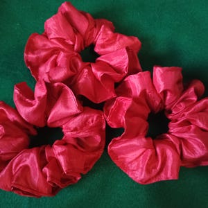 May include: Three red satin scrunchies, each with a gathered, ruffled design. The scrunchies are arranged in a cluster on a green background.