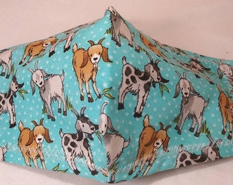 May include: A teal face mask with a repeating pattern of cartoon goats in various poses. The goats are brown, white, and gray, with some featuring black spots. The background is dotted with small white circles. White elastic ear loops are attached.