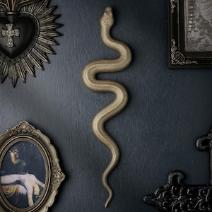 May include: A gold-coloured wall-mounted snake sculpture with a textured, scaled surface. The snake is depicted in a vertical, S-shaped winding position. It is displayed on a dark surface alongside a heart-shaped plaque, a framed picture, and a cross.
