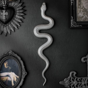 May include: A silver-coloured, textured snake figurine in a winding, S-shaped pose. The snake has a detailed, scaled surface and is positioned vertically, with its head at the top and tail at the bottom, displaying a realistic, serpentine form.