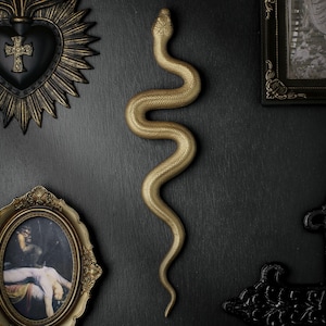 May include: A gold-coloured, textured snake wall sculpture in a vertical, winding position. The snake features a detailed, scaled surface and a distinct head shape, positioned centrally between various framed wall art pieces on a dark surface.