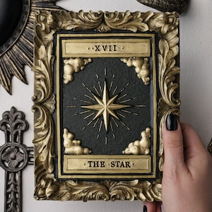 May include: A rectangular wall plaque featuring a black centre panel with a gold embossed eight-pointed star, four gold clouds in the corners, and the Roman numeral XVII at the top. The text THE STAR is at the bottom, encased in an ornate gold frame.