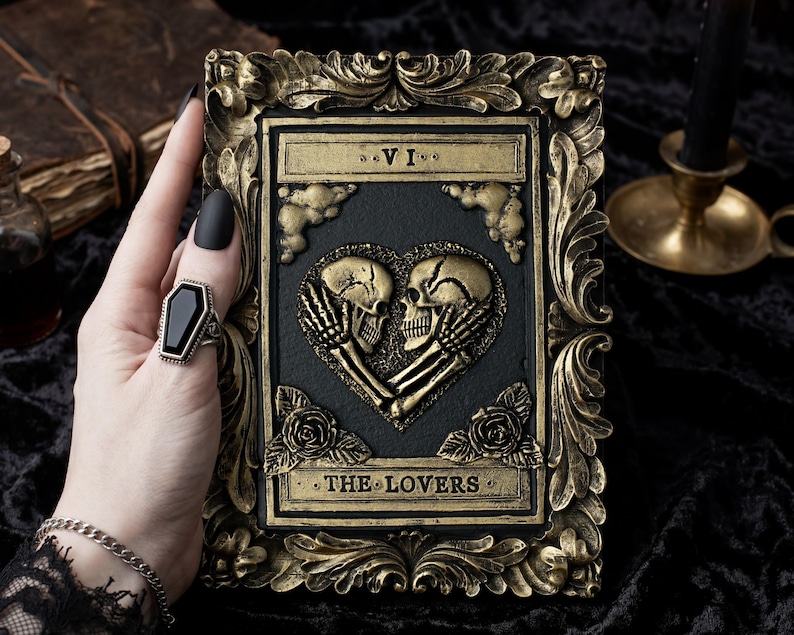 Pu&ograve; includere: Una placca rettangolare con un rilievo di due teschi che formano un cuore, etichettata VI e THE LOVERS. Il pezzo ha un centro nero e una cornice dorata ornata e anticata con accenti floreali. Un anello a forma di bara con una pietra nera.