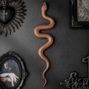 May include: A copper-coloured snake wall decoration with a textured, scaled surface. The snake is positioned in a vertical, S-shaped curve. It is displayed alongside a black heart-shaped plaque with a cross, a framed oval portrait, and a black cross.
