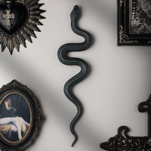 Könnte beinhalten: Eine mattschwarze, an der Wand montierte Schlangenskulptur mit einer texturierten, geschuppten Oberfläche. Die Schlange ist in einer vertikalen, S-förmigen Windung dargestellt, mit einem detaillierten Kopf und einem spitz zulaufenden Schwanz.