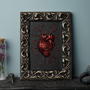 Peut inclure: Un c&oelig;ur anatomique encadr&eacute; avec des veines rouges sur un fond noir. Le cadre est orn&eacute; et dor&eacute;.